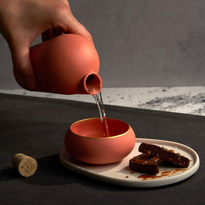 A hand pouring mezcal from a pink carafe into a pink copita sitting on a white plate. Two pieces of chocolate drizzled with goat milk caramel and sprinkled with sea salt sit on the plate. The TUYO branded cork is resting on the ground next to the plate and copita.