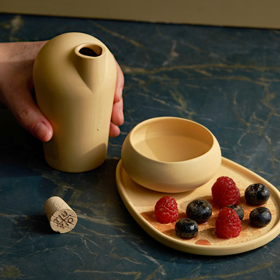 A hand holding a yellow carafe that has just poured mezcal into a matching yellow porcelain copita. In the foreground, a copita rests on a matching yellow plate that holds raspberries and blueberries sprinkled with mezsal salts. The TUYO branded cork is resting on the ground next to the plate and copita.