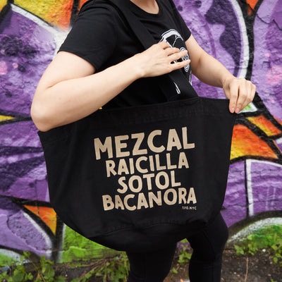 Cotton tote bags black cotton silkscreened with discharge ink on both sides. Text reads: Mezcal, sotol, raicilla, bacanora.