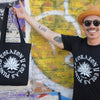 Oscar is wearing a size small black organic cotton tshirt silkscreened with an agave in the center in white ink and the words Puntas, Corazon y Colas and holding a black tote bag with the same design.